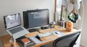 A modern workspace with a laptop and a monitor displaying software, various stationery items, and a plant.