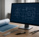 A computer screen displaying blueprints and engineering drawings alongside rolled up architectural plans on a wooden des